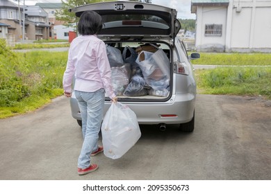 A Young Woman Is Lifting Trash Bag And  Into Trunk Of A Car