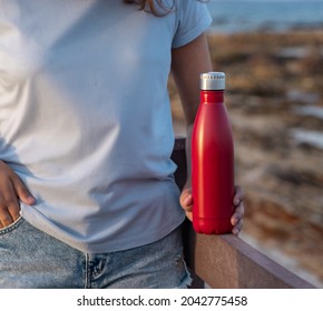 Young Woman In Light Blue T-shirt Holding Red Water Bottle In Hand Close Up, Cropped Shot. Bottle And T-shirt Mockup