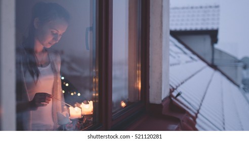 Young Woman Lighting Advent Candles On The Window, Winter Cozy Evening. Winter Holidays Concept. Christmas Traditions. View From Outside The Window. Snow-covered House.