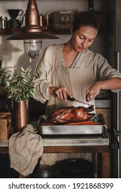 Young Woman In Linen Apron Carving Whole Roasted Duck For Thanksgiving Holiday Or Christmas Eve Party Celebration In Modern Kitchen Interior. Traditional Autumn Or Winter Holiday Comfort Food Concept