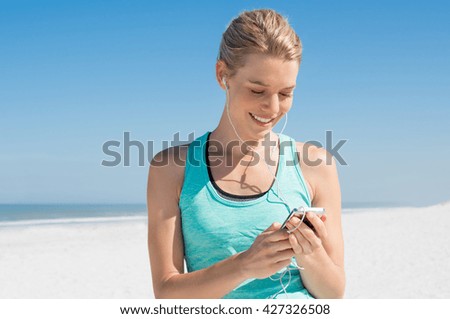 Young woman listening to music during workout. Runner woman checking her song with mp3 player while resting from routine exercise. Happy young jogger listening to music at beach.
