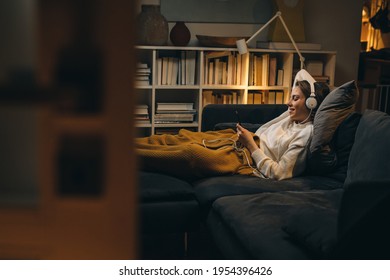 young woman listening to a music on headset. she is relaxing on sofa at her living room. evening scene - Powered by Shutterstock