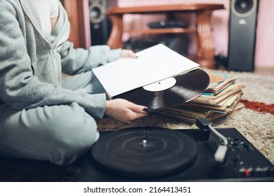 Young Woman Listening To Music From Vinyl Record Player. Retro And Vintage Music Style. Girl Holding Analog Record Album Sitting In Room At Home. Female Enjoying Music From Old Record Collection