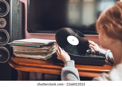 Young Woman Listening To Music From Vinyl Record Player. Retro And Vintage Music Style. Girl Holding Analog Record Album Sitting In Room At Home. Female Enjoying Music From Old Record Collection