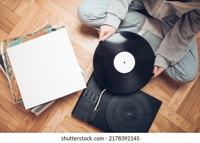 Young Woman Listening To Music From Vinyl Record Player. Retro And Vintage Music Style. Girl Holding Analog Record Album Sitting In Room At Home. Female Enjoying Music From Old Record Collection