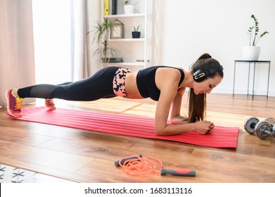 A Young Woman Listens Music In Headphones And Does Sports At Home, She Does An Exercise Plank. Healthy Lifestyle Concept