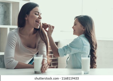 Young Woman And Little Girl Eating Muffins. Adorable Daughter Feeding Beautifull Smiling Mother With Muffin. Happy Mother And Child Sitting At Table Drinking Milk Together At Home