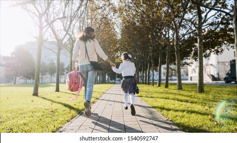 Young Woman And Little Girl In School Uniform Are Holding Hands And Running Along The Trees In The Park At Sunny Autumn Weather. Family Late For School Concept, Back View.