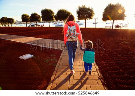 Young woman and little toddler boy walking through a park to the sea. Mother in orange jacket with backpack and small son go to the quay with fish boats and yachts. Beautiful green trees on sunset.