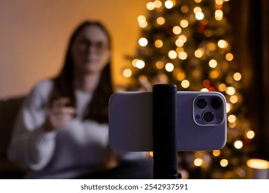 Young woman live streaming or recording holiday video message on smartphone, with Christmas tree lights creating cozy festive background ambiance  - Powered by Shutterstock