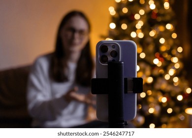 Young woman live streaming or recording holiday video on smartphone with Christmas tree in background, cozy festive ambiance at home - Powered by Shutterstock