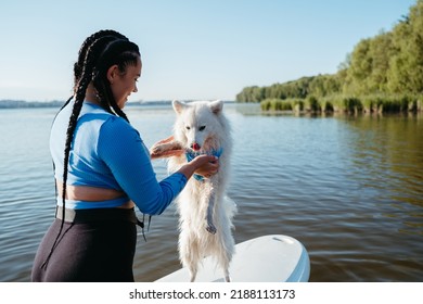 Young Woman With Locs Holding Her Pet Japanese Spitz On The Lake, Dog Standing On Sup Board On His Back Paws