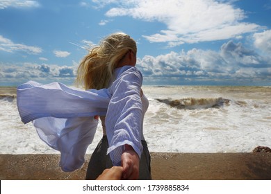 Young woman with long blond hair  looks at stormy waves and holds man's hand. Follow me concept. Strong waves, heavy clouds on stormy weather. The Black sea coast in summer stormy day. Love story - Powered by Shutterstock