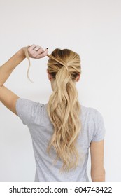 A Young Woman With Long Blond Wavy Hair Tying A Ponytail Back View