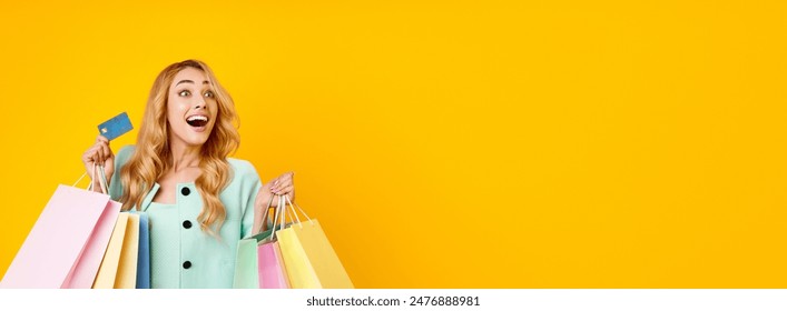 A young woman with long blonde hair is standing in front of a yellow background. She is holding multiple shopping bags and a credit card. She is smiling and looking to the copy space, panorama - Powered by Shutterstock