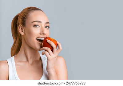 A young woman with long blonde hair tied back in a ponytail smiles as she bites into a red apple. She is wearing a white tank top and is standing in front of a gray background, copy space - Powered by Shutterstock