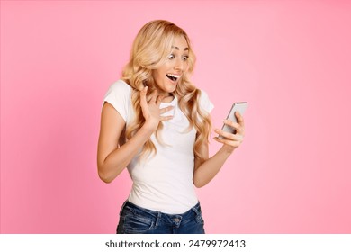 A young woman with long blonde hair is shown in front of a pink background. She is wearing a white t-shirt and blue jeans and is looking at her phone with an excited expression - Powered by Shutterstock