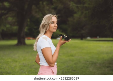 A young woman with long blonde hair is standing outdoors, holding a smartphone to her ear. She is wearing a white top and pink pants, looking thoughtfully into the distance. The background features gr - Powered by Shutterstock