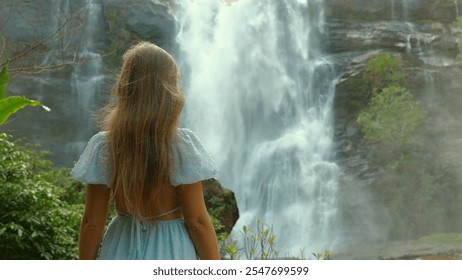 Young woman with long blonde hair in light blue dress stands in front of majestic waterfall, surrounded by lush greenery, admiring natural beauty and serenity of scene. Nature and Tranquility - Powered by Shutterstock