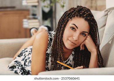 Young woman with long braids writes in a notebook on her sofa, looking relaxed and content in her cozy living room. She smiles, lost in thought and creativity, embracing her natural beauty - Powered by Shutterstock