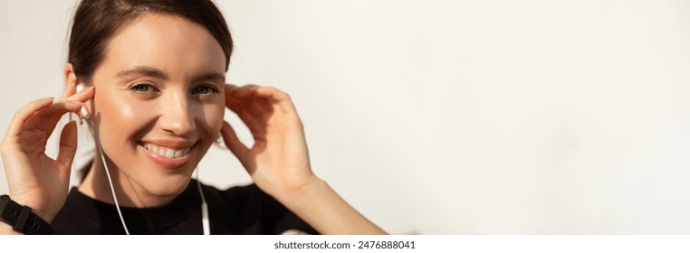 A young woman with long brown hair smiles while adjusting her white earbuds. Her skin is glowing, and she appears happy and relaxed, copy space - Powered by Shutterstock