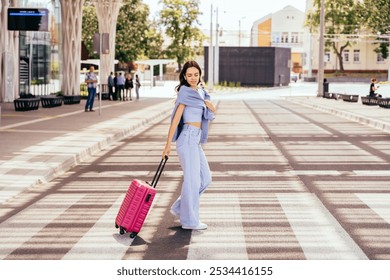 Young woman with long brown hair stands at urban street crossing pulling pink suitcase, dressed in blue jeans and top. Sunny day, summer travel vibe. Pavement and modern architecture in background. - Powered by Shutterstock