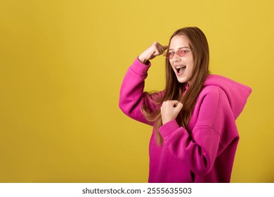 A young woman with long brown hair, wearing a pink hoodie with the hood up and pink sunglasses, smiles broadly and joyfully makes a yes gesture with her fists. - Powered by Shutterstock