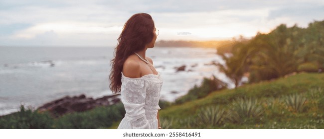 A young woman with long brown hair gazes at the sunset over the ocean, standing on a lush tropical coastline. She wears an off-shoulder white dress, capturing a serene moment of natural beauty and - Powered by Shutterstock