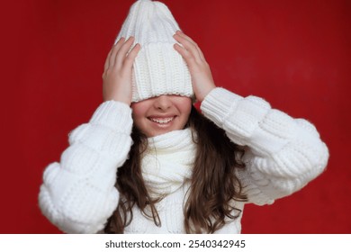 a young woman with long chestnut hair is playfully obscured by a cozy white knitted hat that rests over her eyes. She holds a soft, textured scarf in her hands against a vivid red background.. - Powered by Shutterstock