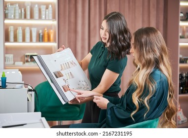 Young woman with long curly hair sitting in salon chair and pointing at hair sample while choosing hair dye with hairdresser. Female hairstylist holding holder with hair samples. - Powered by Shutterstock