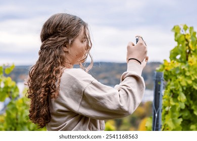 A young woman with long curly hair capturing scenic vineyard landscape views with her smartphone. Teenager blogger girl photographing autumn nature with her phone. Copy space - Powered by Shutterstock