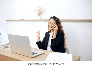 A young woman with long, curly hair smiles during a phone call and takes notes at her tidy desk. The bright office features light wood accents and minimal decor, creating an inviting workspace. - Powered by Shutterstock