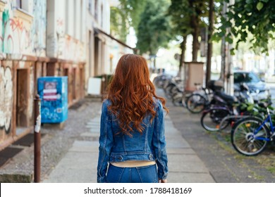 Young woman with long curly red hair wearing denims walking away down an urban street