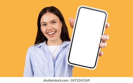 A young woman with long dark hair smiles broadly as she holds a smartphone with a blank white screen towards the camera. She is standing against a bright yellow background. - Powered by Shutterstock