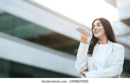 A young woman with long dark hair is standing outdoors in an urban environment and holding a smartphone to her mouth as she uses a voice assistant, copy space - Powered by Shutterstock