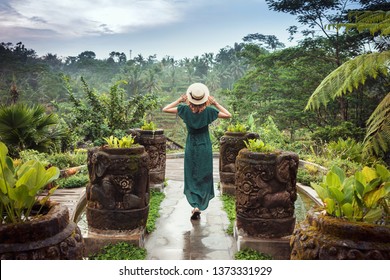 A young woman in a long dress of emerald color and wearing a hat is standing with her back against the background of a green tropical jungle, Ubud, Bali. Girl in a romantic image among the plants - Powered by Shutterstock