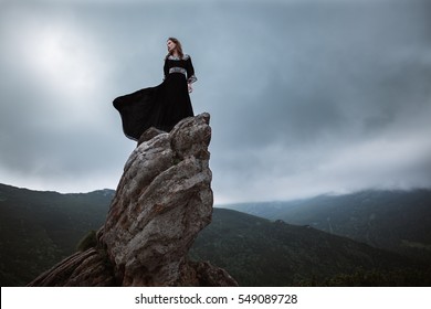 Young woman in long dress enjoying nature on the mountains. The girl in black dress with long flying train stands on the top of the rock