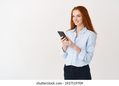 Young Woman With Long Ginger Hair And Blue Eyes Reading Message On Phone, Smiling And Looking Happy While Using Smartphone, White Background.