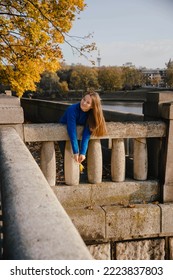 Young Woman With Long Hair In Blue Sweater And Maple Leaf In Her Hand Bent Over The Stone Banister Against The Backdrop Of Yellow Tree And A River. 