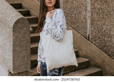 Young woman with long hair holding tote bag for mockup design. The woman is on stairs, wearing a casual outfit holding white cloth bag. The tote bag is white for mockup, and the woman appears relaxed.