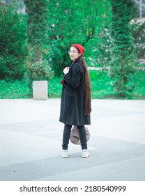 A Young Woman With Long Hair And A Red Hat Wearing Glasses And Dark Coat With A Red Scarf In The Park Looking Backwards Against Trees On A Pedestrian Road. Bright Summer Evening