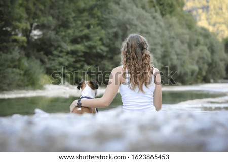 Young woman with long hair sits together with a small terrier dog on a river bank, rear view, lights bokeh
