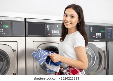 A young woman with long hair smiles while holding clothes in a laundromat. She stands by washing machines, actively engaged in doing her laundry and enjoying the experience. - Powered by Shutterstock