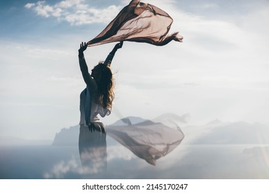 Young woman with long hair stands on coastline rocky cliff edge and holds shawl waved by strong wind against sea under morning mist