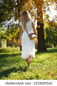 Young woman with long hair, in white summer dress walking bare foot on green park meadow, view from behind - Shutterstock ID 2115029162
