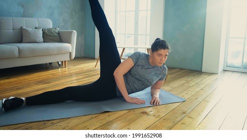 Young Woman With Long Ponytail In Sportswear Does Supine Side Leg Raises Lying On Mat In Spacious Living Room