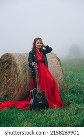 Young Woman In Long Red Dress And Leather Jacket With Guitar Standing Near Haystack In Mist Field At Twilight Time. Creative Millennial Women Hobbies And Interests