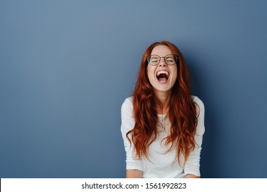 Young woman with long red hair and glasses enjoying a loud guffaw or belly laugh over a blue studio background with copy space - Shutterstock ID 1561992823