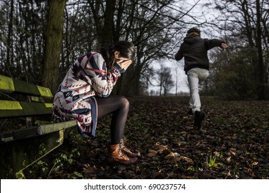 A Young Woman With A Long Sweater Sitting On A Bench In Dark Forest.She Covers Her Eyes While Crying.A Man Is Running Away.A Concept About Being Attacked,hurt,harrased Or Raped.Hard Flash Light Image.