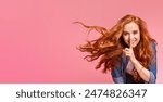 A young woman with long, vibrant red hair flows to the side as she places one finger to her lips, signaling for silence on pink background, copy space